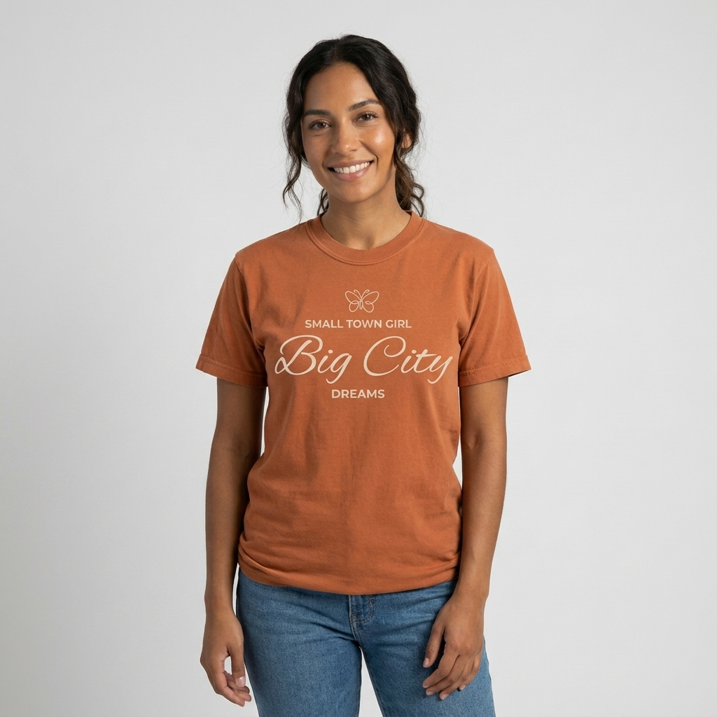 A woman wearing a burnt orange graphic t-shirt with the text 'Small Town Girl Big City Dreams' in white, paired with blue jeans, against a light gray studio background.