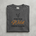 A folded dark gray t-shirt with a graphic print that reads 'BORN TO BE Wild FORCED TO WORK' in black and orange lettering, with a small black wildflower illustration above. The shirt is laid flat on a textured gray background.