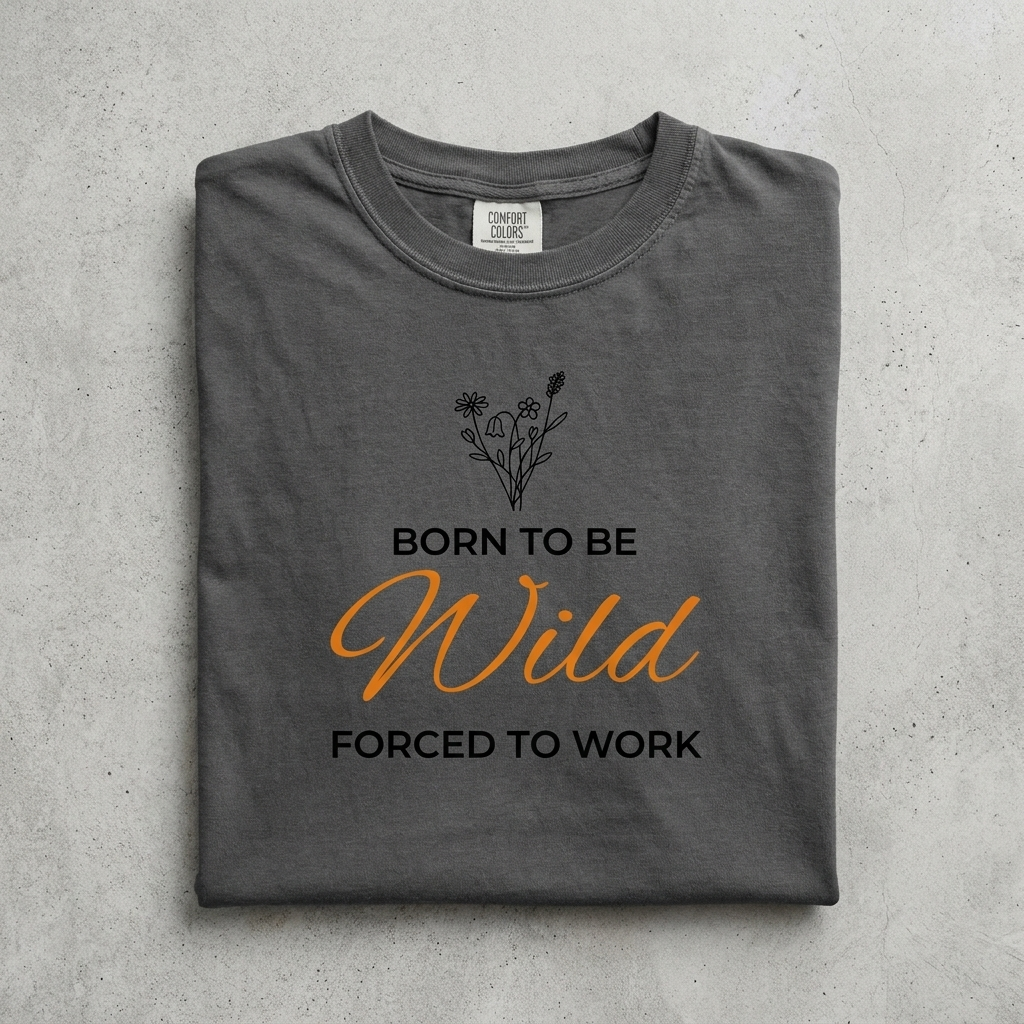 A folded dark gray t-shirt with a graphic print that reads 'BORN TO BE Wild FORCED TO WORK' in black and orange lettering, with a small black wildflower illustration above. The shirt is laid flat on a textured gray background.