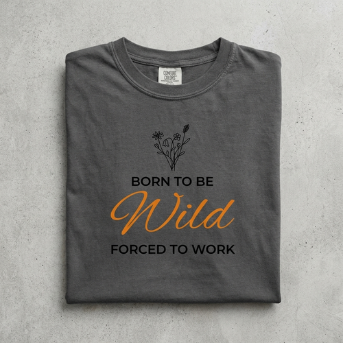 A folded dark gray t-shirt with a graphic print that reads 'BORN TO BE Wild FORCED TO WORK' in black and orange lettering, with a small black wildflower illustration above. The shirt is laid flat on a textured gray background.