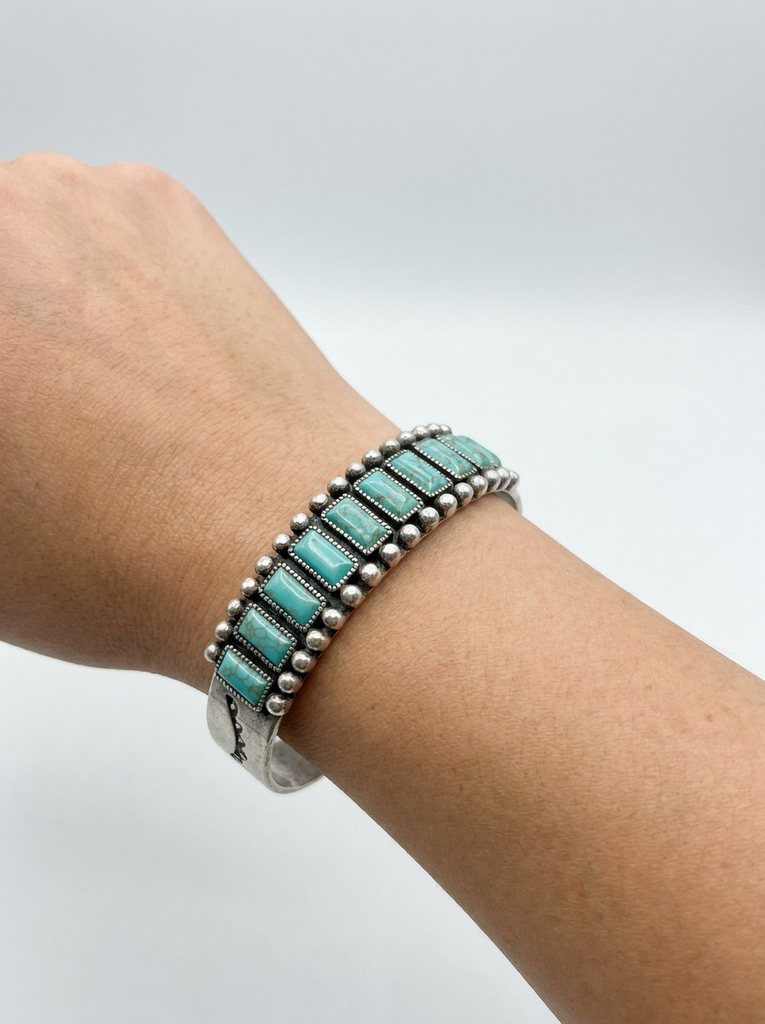 A sterling silver cuff bracelet with rectangular turquoise stones, worn on a person's wrist against a neutral light gray background.