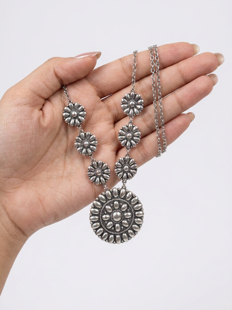 A silver-toned bohemian-style necklace with a large circular medallion and five smaller flower-shaped links, held in a person's hand against a plain light gray background.