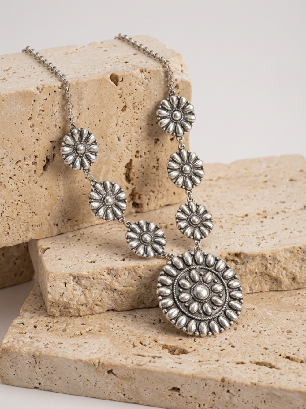 An antique silver-toned statement necklace with floral disc motifs and a large circular medallion pendant, displayed on stacked beige travertine stones against a light neutral background.