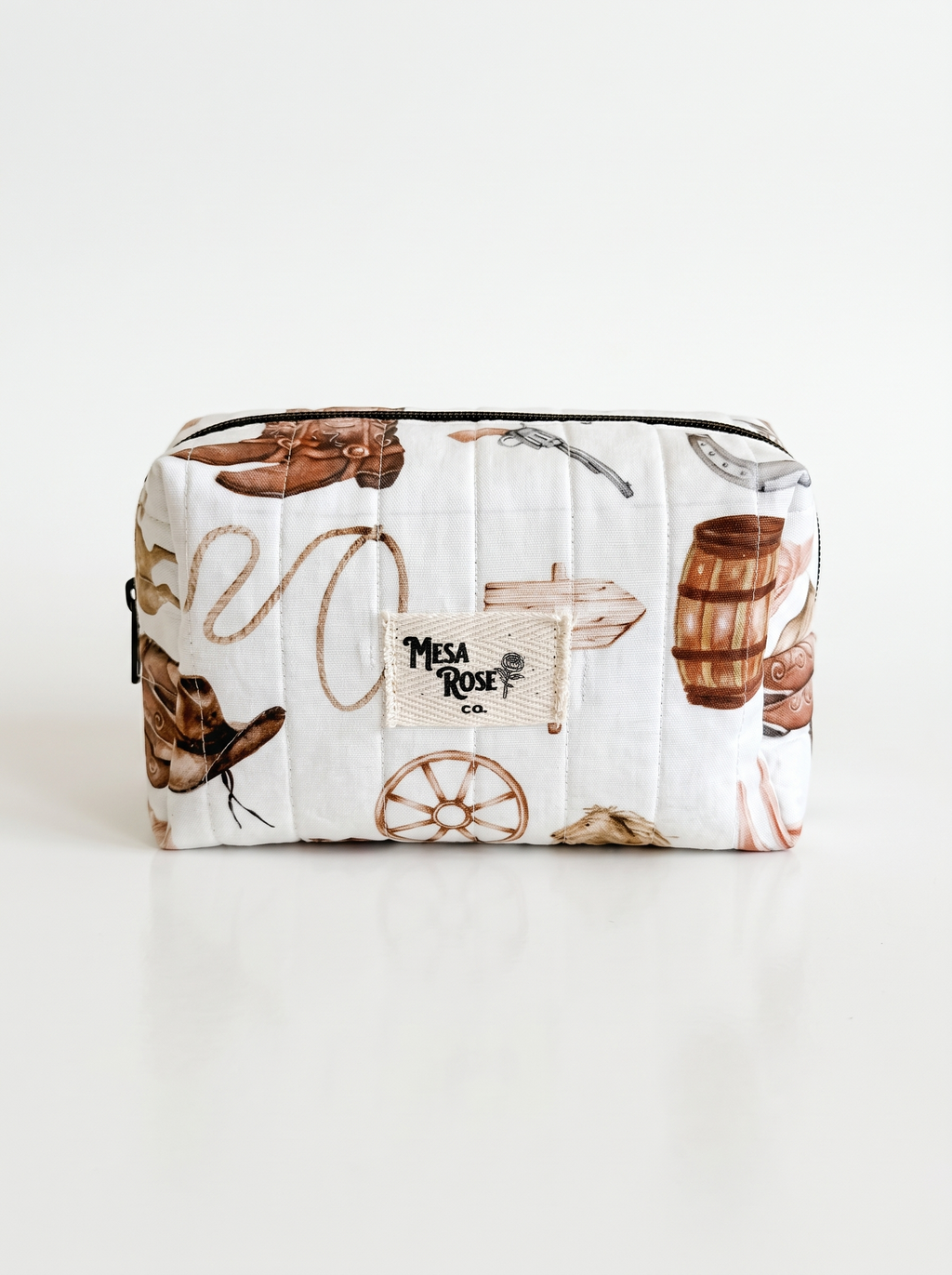 A white quilted cosmetic pouch with a western-themed print featuring brown cowboy boots, a lasso, a wagon wheel, a wooden barrel, and a central patch that reads 'Mesa Rose Co'.
