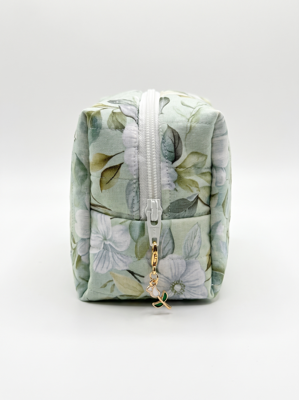 A small rectangular zippered pouch with a light sage green floral print, featuring a white zipper and a small gold charm, photographed on a seamless light gray background.