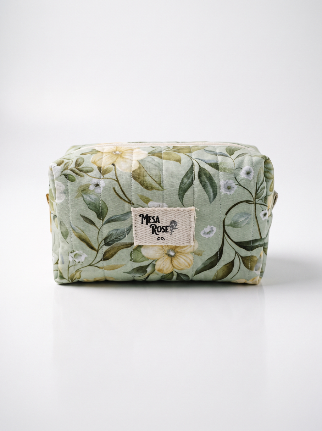 A rectangular quilted cosmetic pouch with a light sage green floral print, featuring a central beige fabric label that reads 'Mesa Rose Co.' on a clean white background.