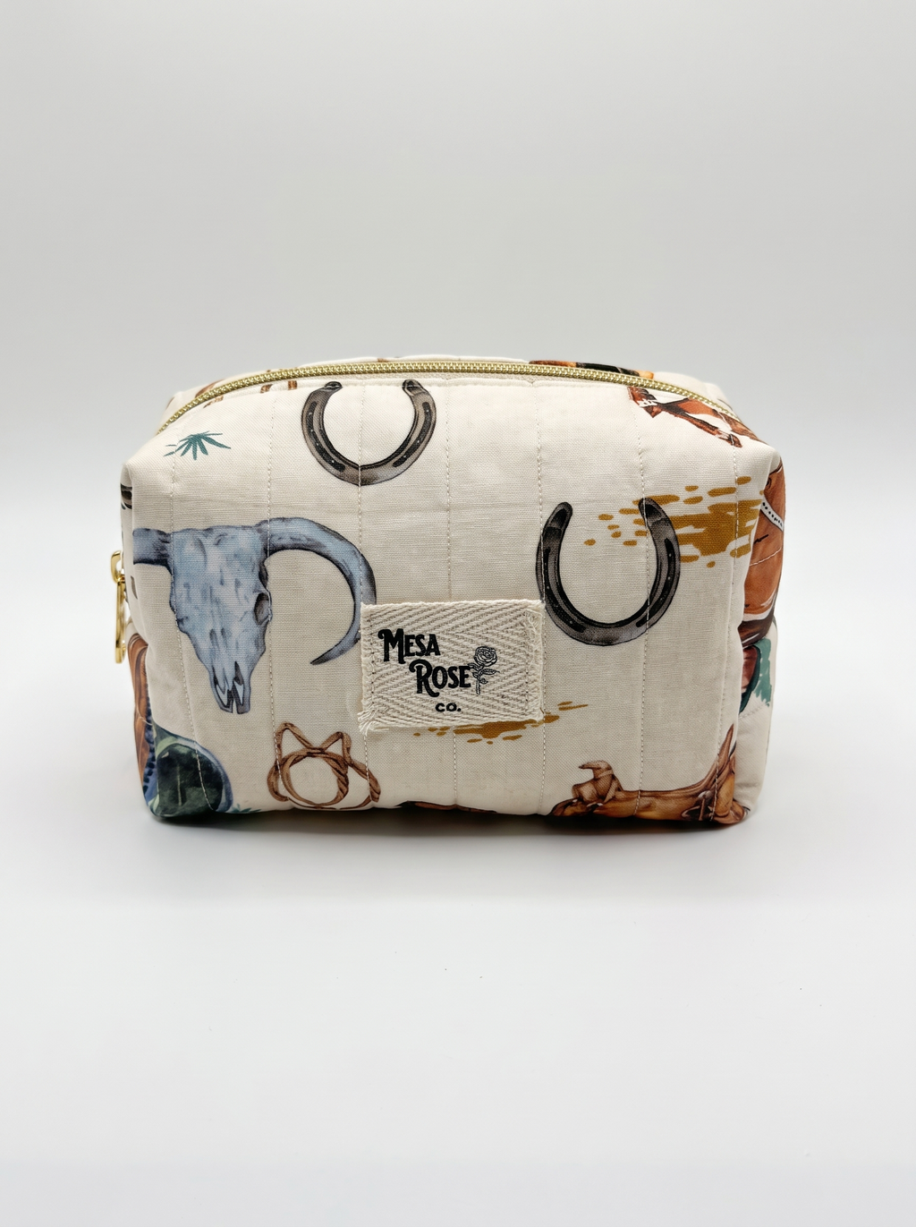 A quilted cosmetic pouch by Mesa Rose Co. featuring a western-themed print with horseshoes, a blue skull, and brown boots on an off-white background, with a gold zipper.