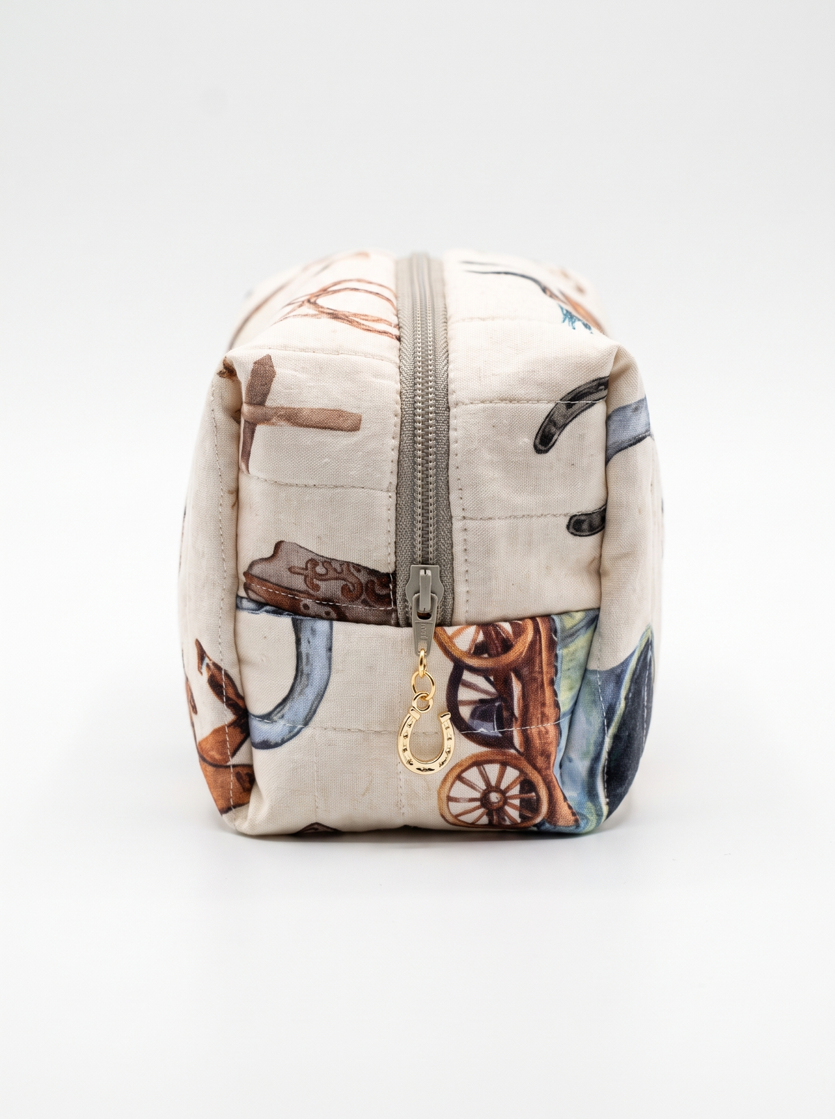 A small zippered cosmetic pouch with a western-themed print featuring horseshoes, a wooden wagon, and a cowboy boot on a light beige background, shown against a plain white studio background.