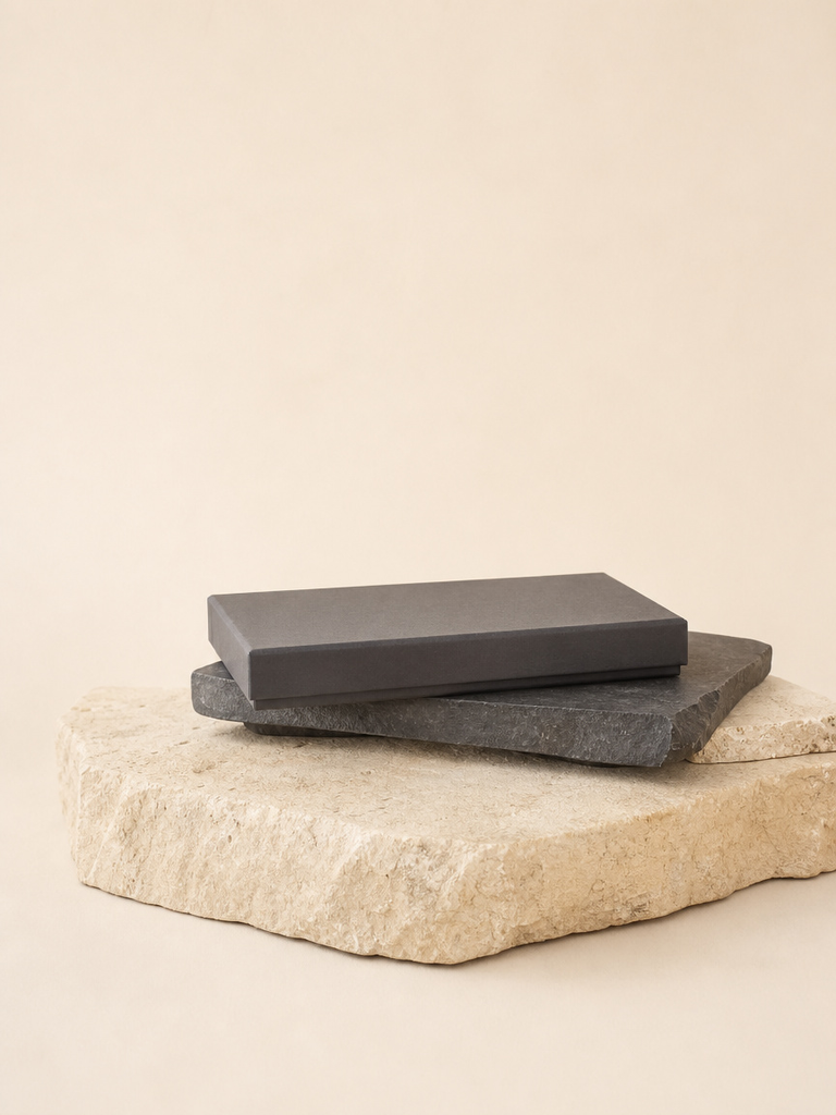 Two rectangular dark gray stone blocks stacked on a larger natural beige stone slab, photographed against a warm cream studio background.