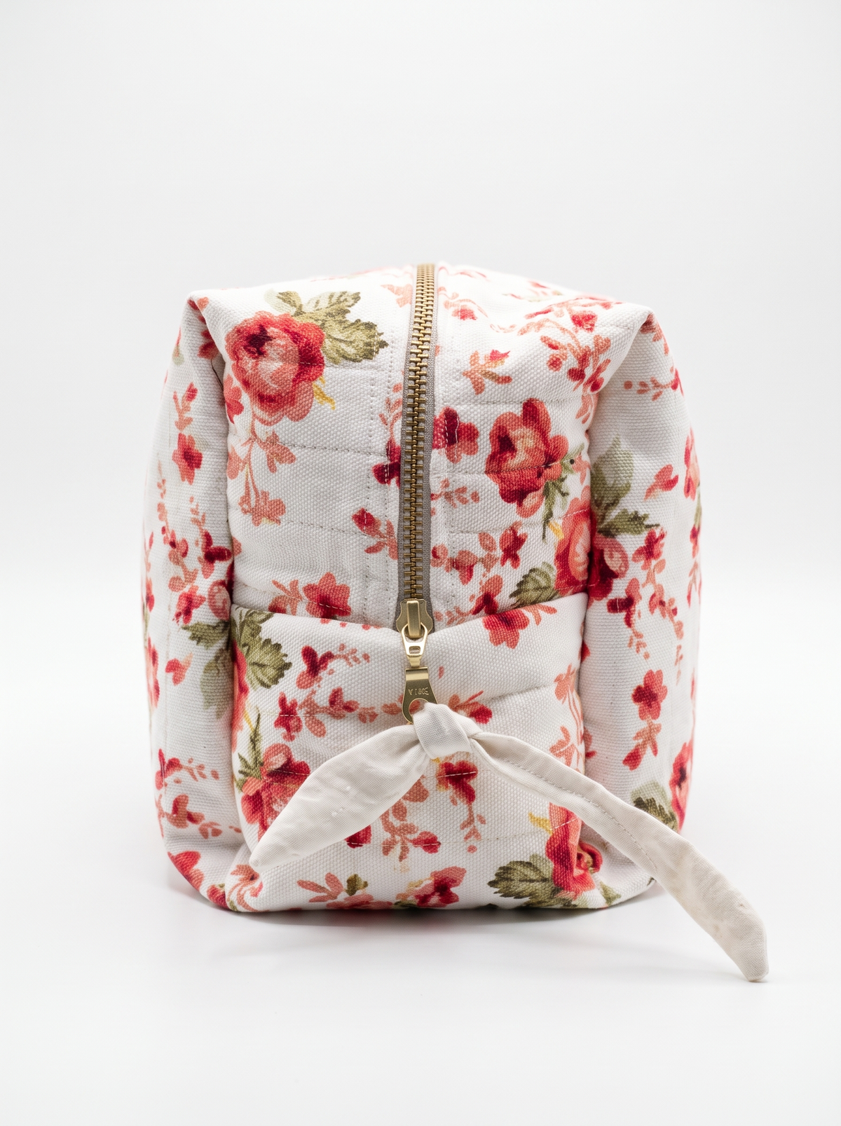 A small quilted fabric pouch with a red and green floral print on a white background, featuring a gold zipper and a white fabric tie closure.
