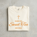 A folded cream-colored t-shirt with orange text and a cross graphic reading 'RAISED ON Sweet Tea AND JESUS', displayed on a textured gray concrete surface.