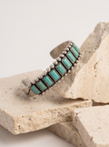 A silver cuff bracelet with rectangular turquoise stones, displayed on stacked natural stone blocks against a light neutral background.