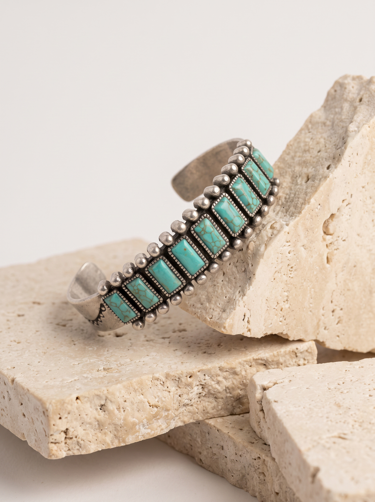 A silver cuff bracelet with rectangular turquoise stones, displayed on stacked natural stone blocks against a light neutral background.